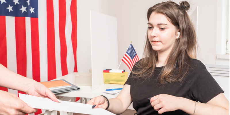 A young woman is seated in a US embassy office, receiving her visa document. The USA flag and files are beside her.