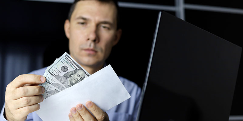 Man in office clothes pulls US dollars money out of an envelope as he sits at his laptop
