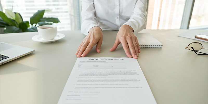 A female HR professional in a white shirt offering an employment agreement for reading labor terms and work conditions. A coffee cup, spec notepad, and mobile phone are placed on the table.