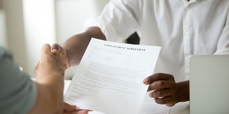 Mid close up shot of two businesspeople shaking hands and a person holding a form labeled "Employee Agreement"