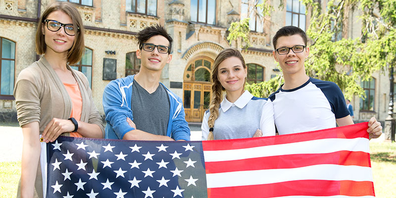 A group of students holding the flag of the USA stands on the campus of a university