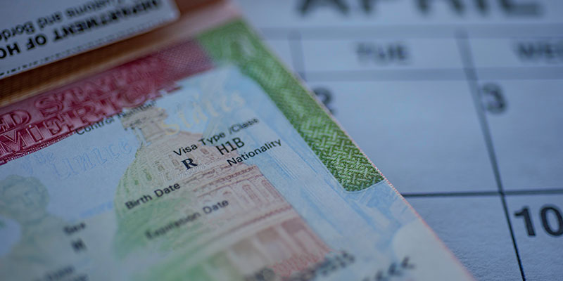 A close-up of the H1B visa page and stamp in a passport with a blurred April calendar in the background